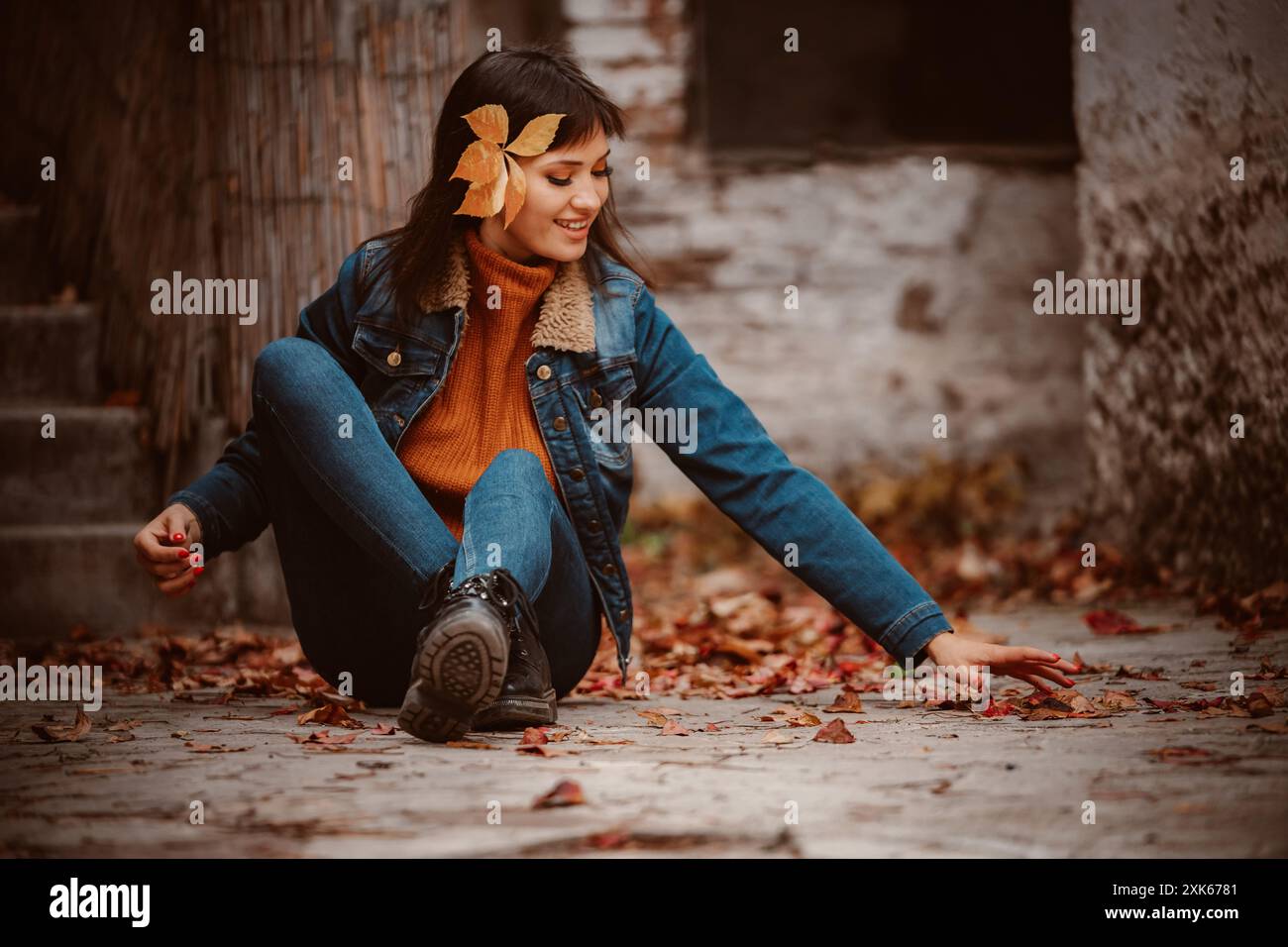 A young woman with a leaf in her hair sits on a stone path surrounded by fallen leaves. Stock Photo