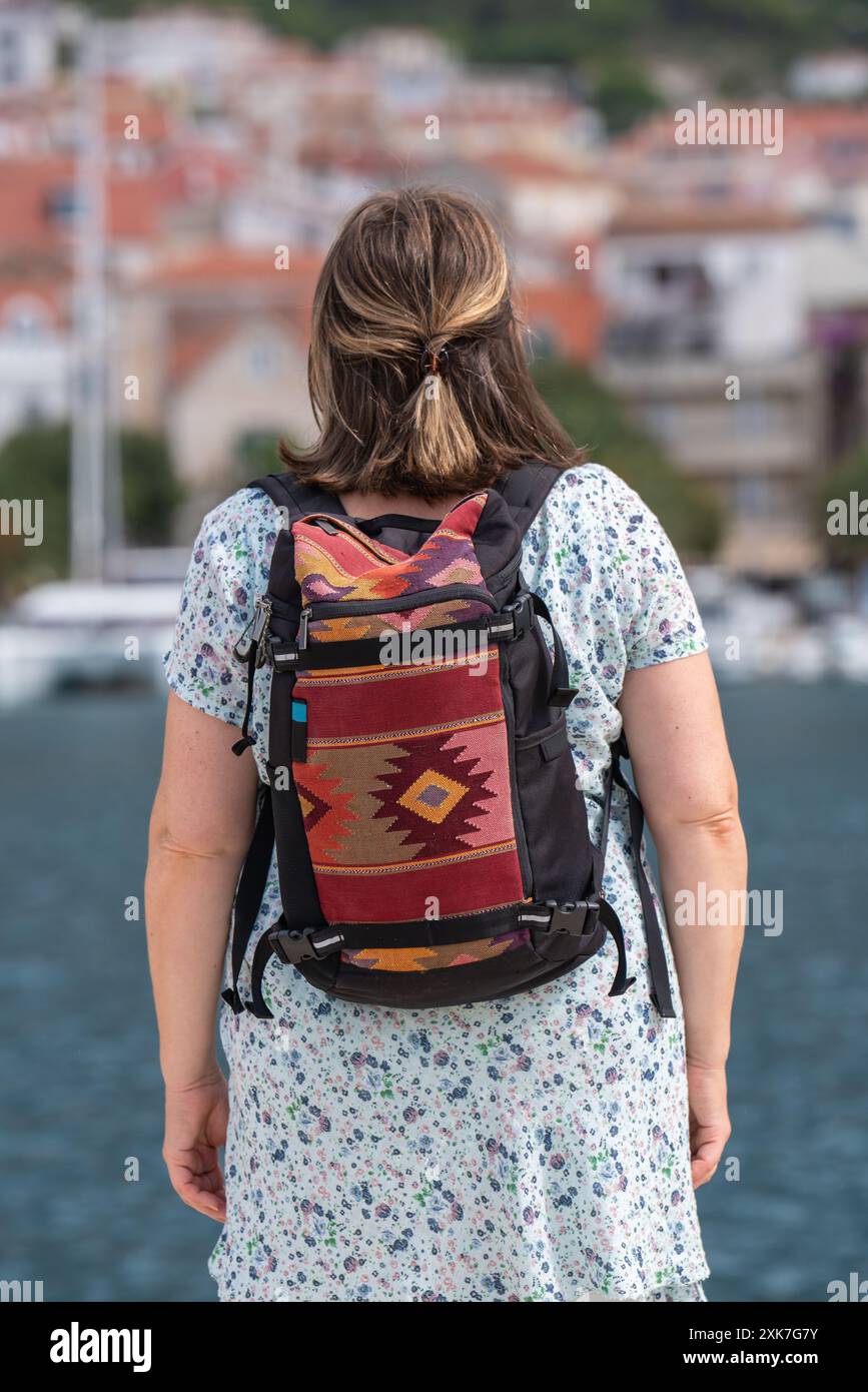 A middle-aged woman travels the world. Stands on the back. On the back is a backpack with an ornament. He looks at the sea and the city in front of hi Stock Photo