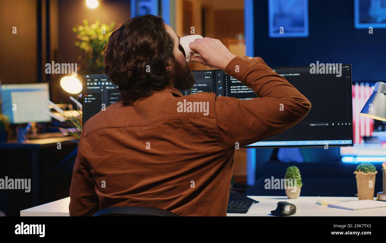 Software engineer writes binary code scripts on computer and drinks coffee. IT specialist in home office enjoying hot beverage while using programming to safeguard consumer data, camera A Stock Photo