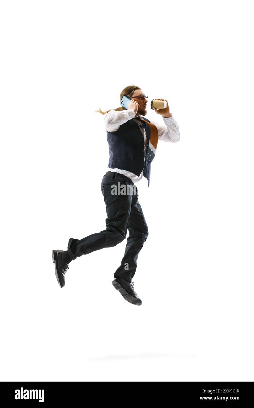 Man in business attire leaps mid-air while multitasking, holding phone to his ear and coffee cup in other hand against white background. Movement. Stock Photo