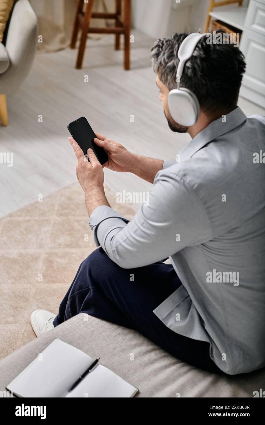 A bearded man wearing headphones sits on a couch, using his smartphone Stock Photo