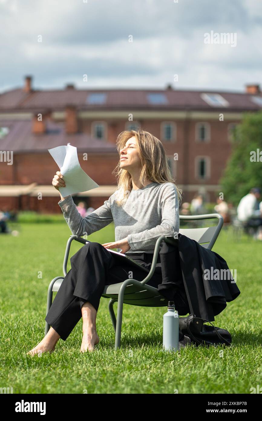 Urban heat island in city. Exhausted woman squints in bright sun, fans herself with papers in park Stock Photo