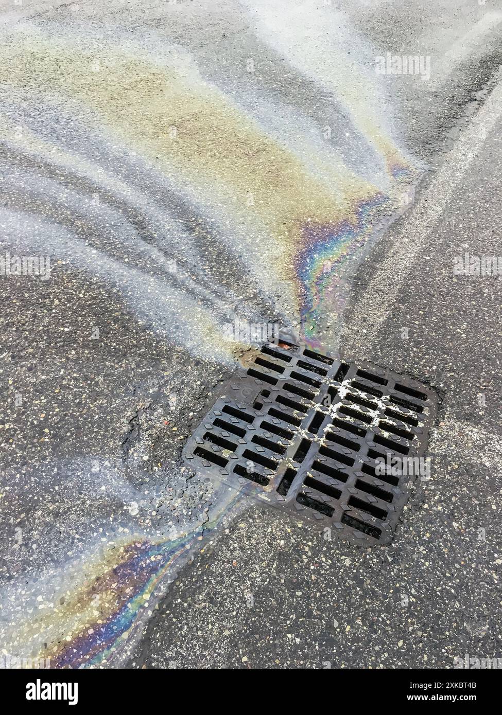 Puddle of motor oil and gasoline on the asphalt road seeps into a nearby sewer grate. Oil stain on the asphalt surface seeps into the storm drainage s Stock Photo