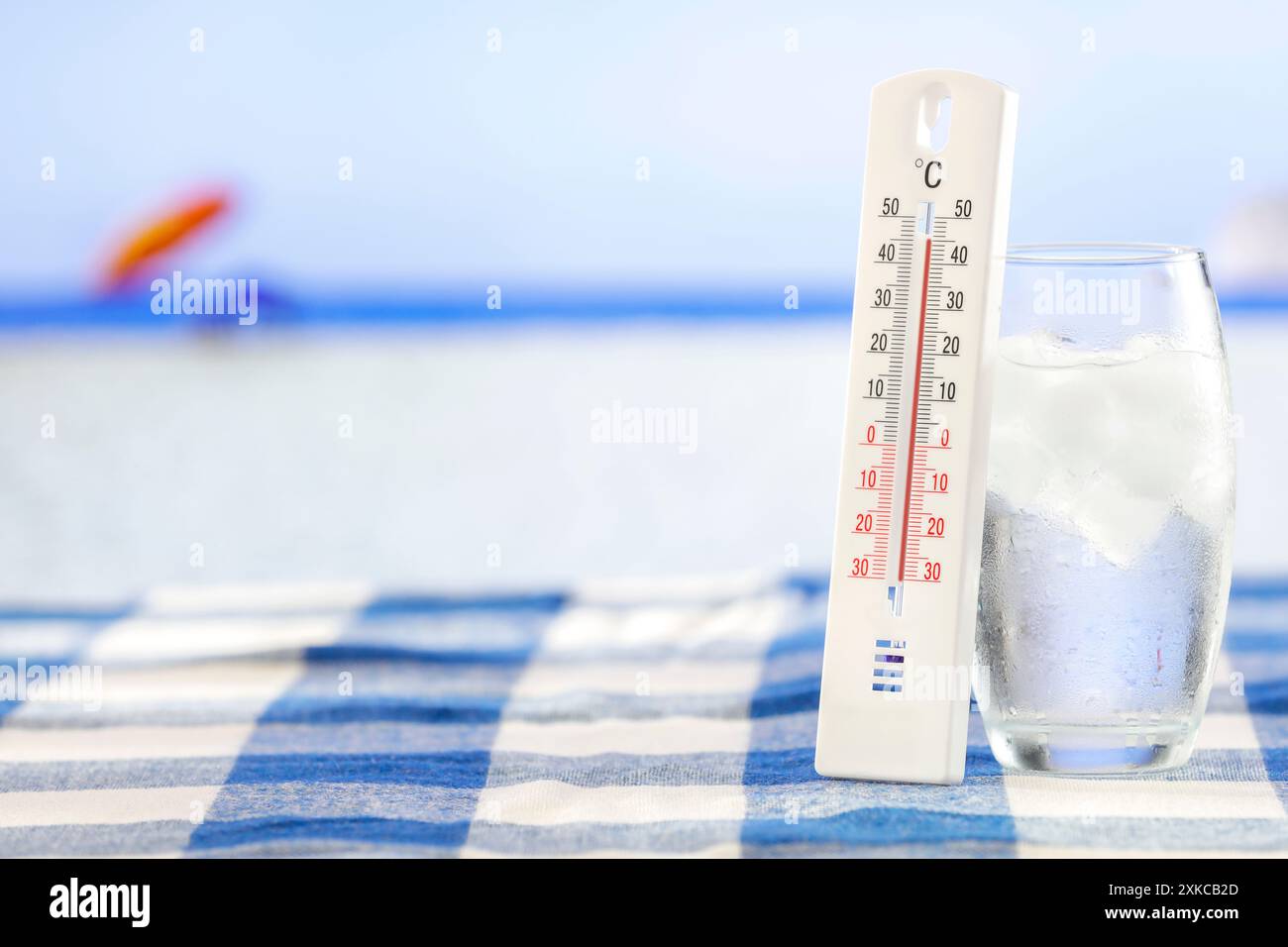 Glass with cold water with ice cubes and thermometer and beach and sea background. Summer cool drink on hot days. Copy space for products. Stock Photo