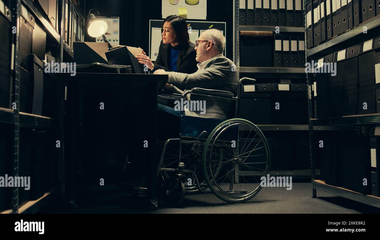 Inspector consulting with criminal expert on archived materials and autopsy records, examining folders and old cases to find connections. Private investigation in disability friendly space. Camera B. Stock Photo