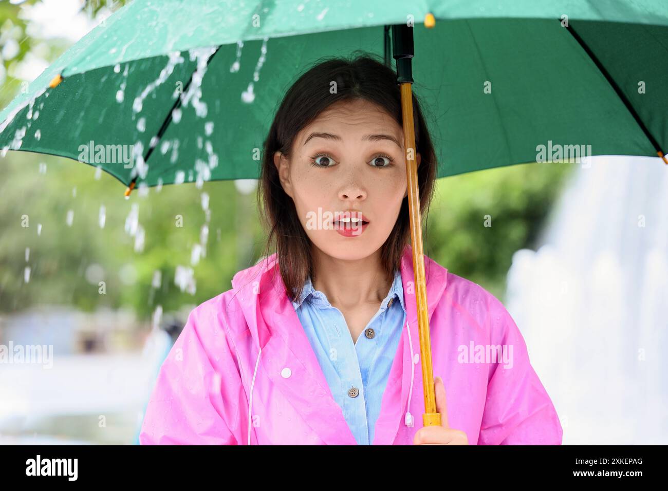 Portrait of surprised young Asian woman man in raincoat holding umbrella outdoors on rainy day Stock Photo