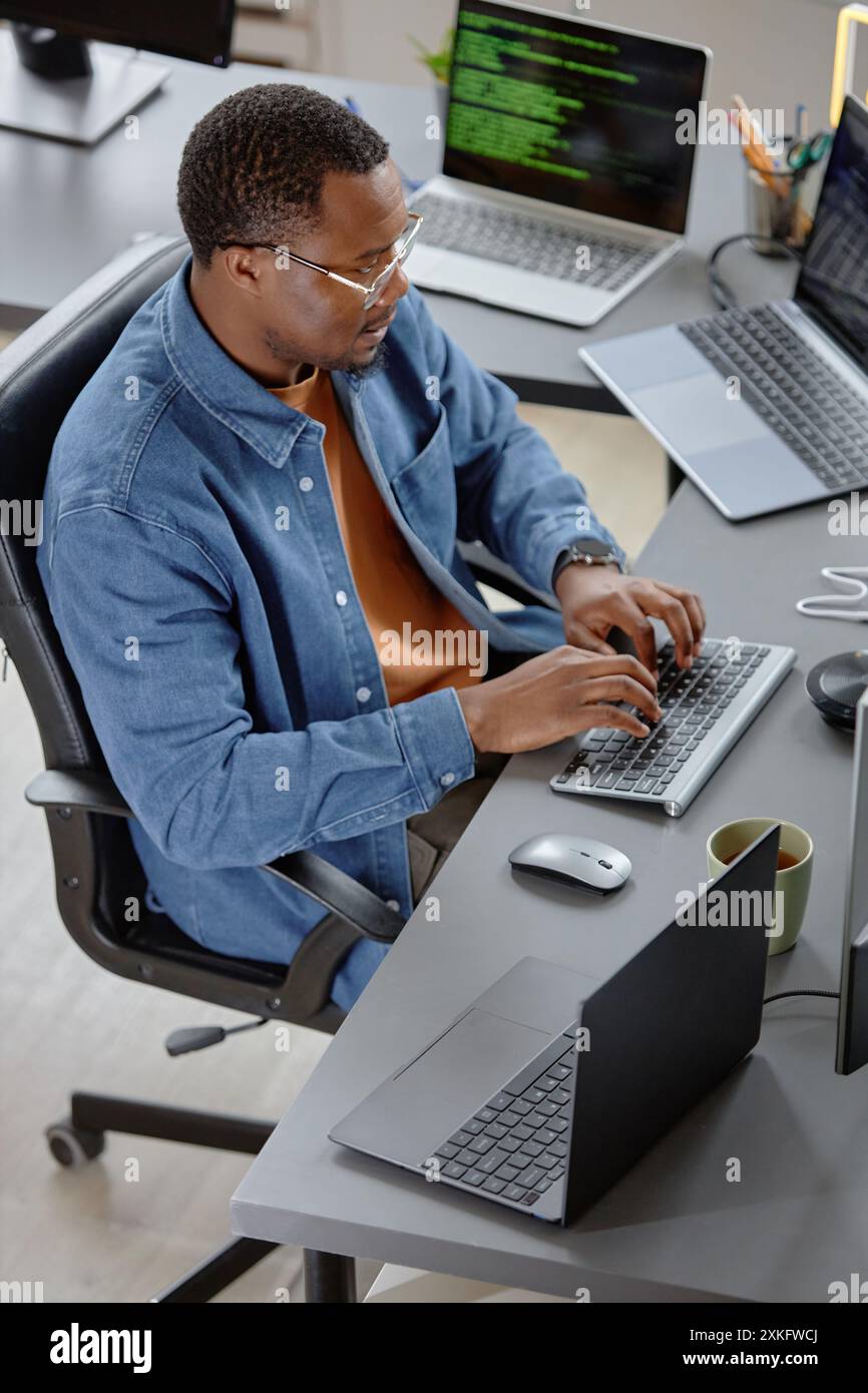 Vertical high-angle shot of concentrated male African American software developer writing program code fixing errors while sitting at working desk with laptops in office Stock Photo