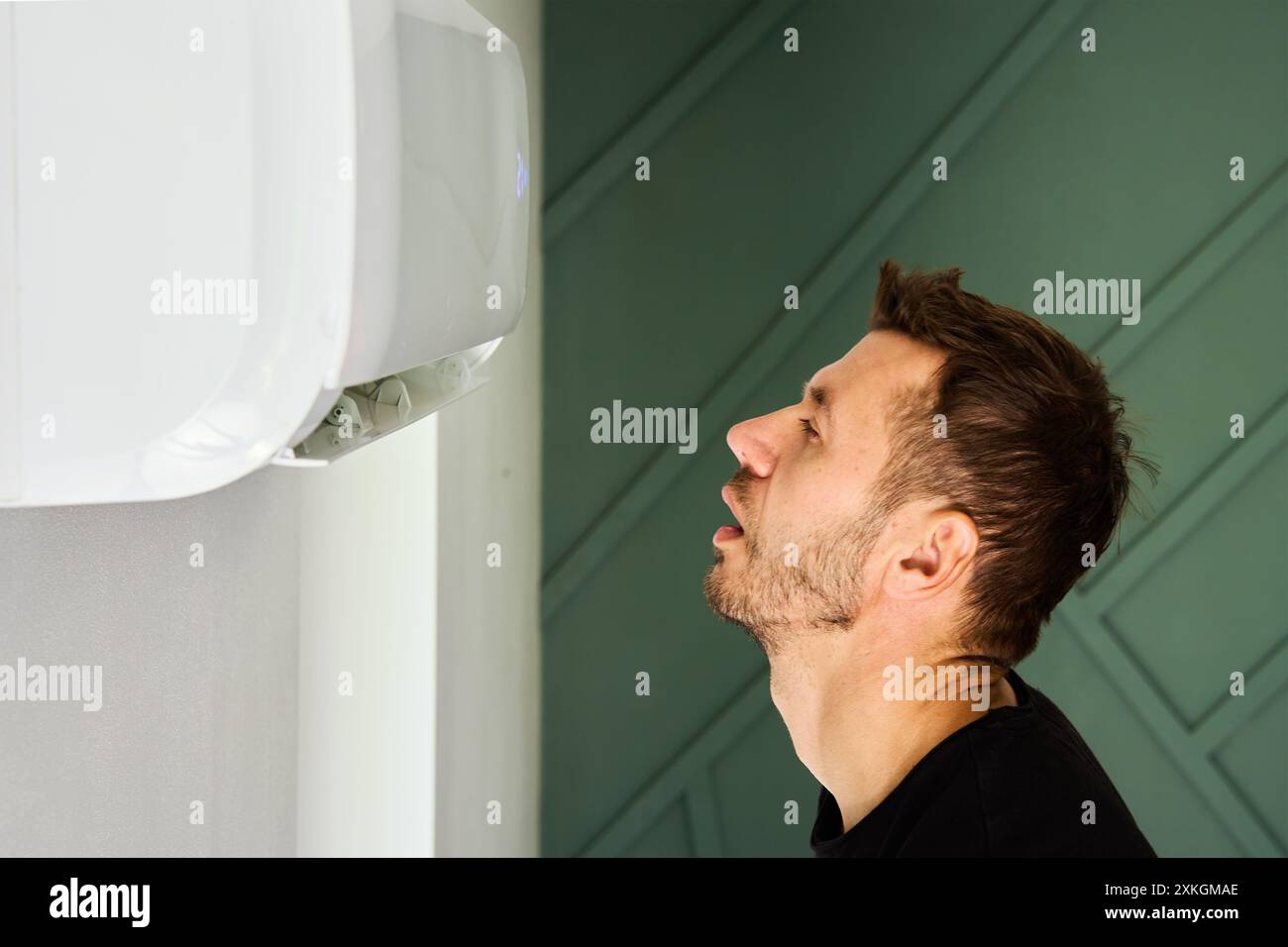 Man stands in living room by air conditioner and enjoys cool breeze. Person escapes from exhausting heat under air conditioner Stock Photo