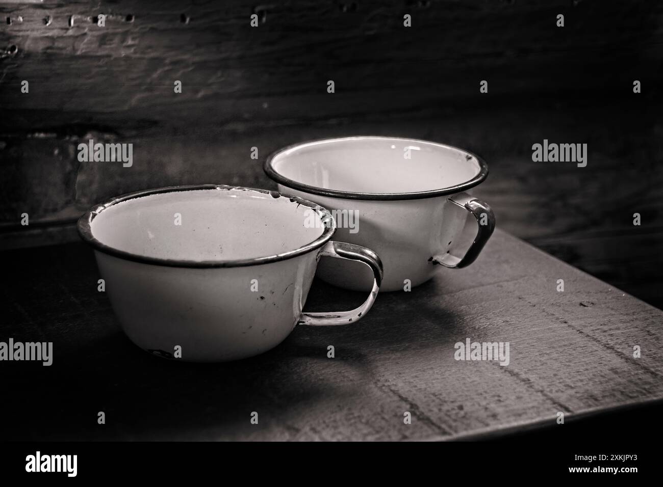 Black and white image; two metal cups on a small wood table inside a vintage log cabin. Soft lighting enters from the upper left illuminating subject. Stock Photo