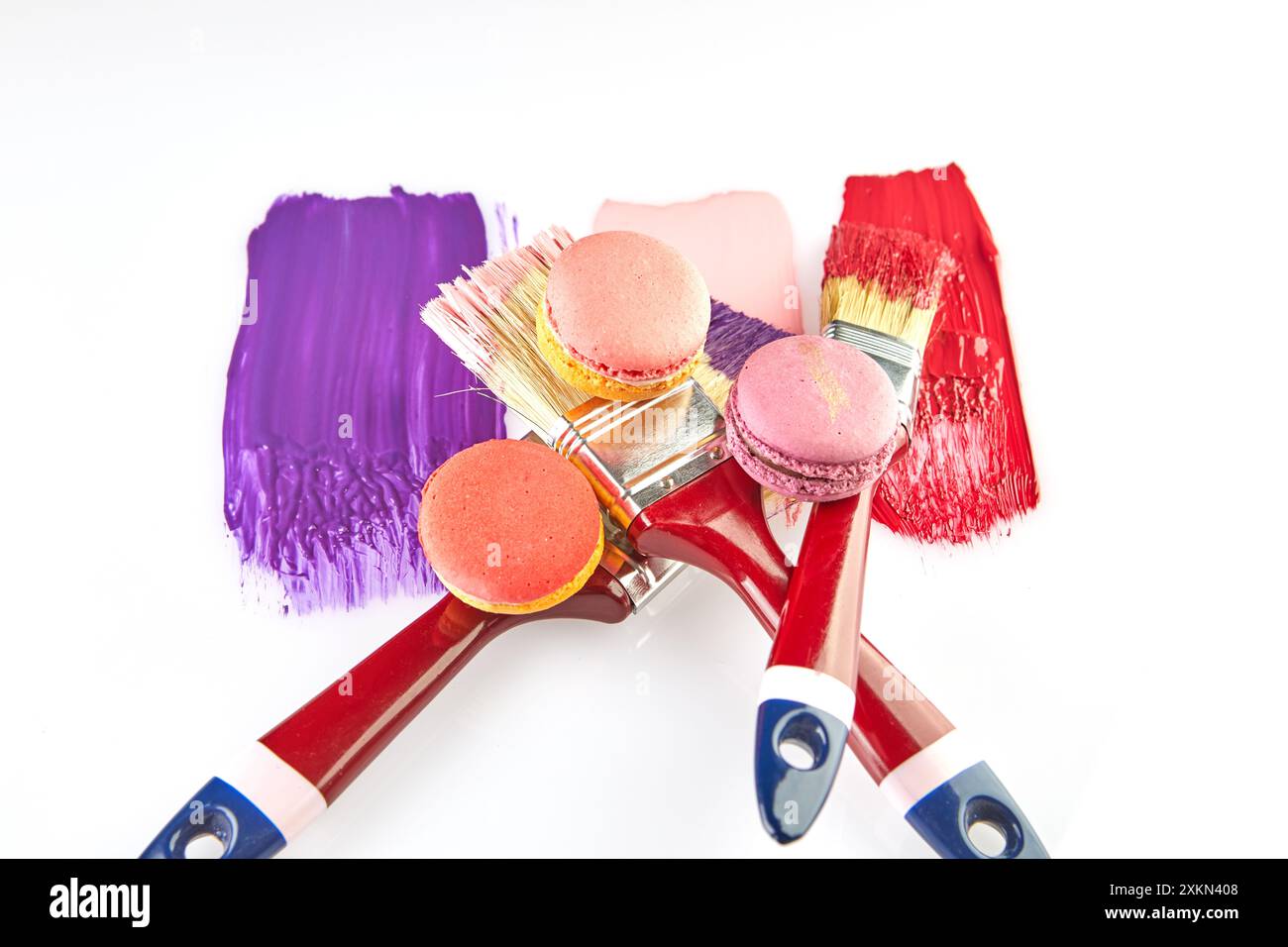 Three paintbrushes with red handles, each dipped in colorful paint, with matching macarons on a white background. Stock Photo