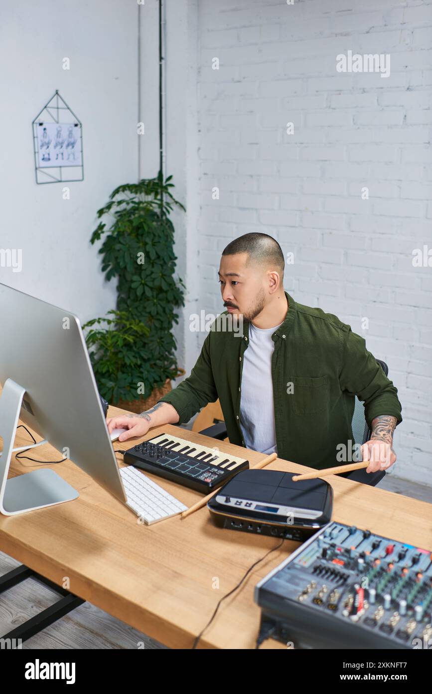 An Asian man in casual attire sits at his desk in his studio, playing various musical instruments. Stock Photo