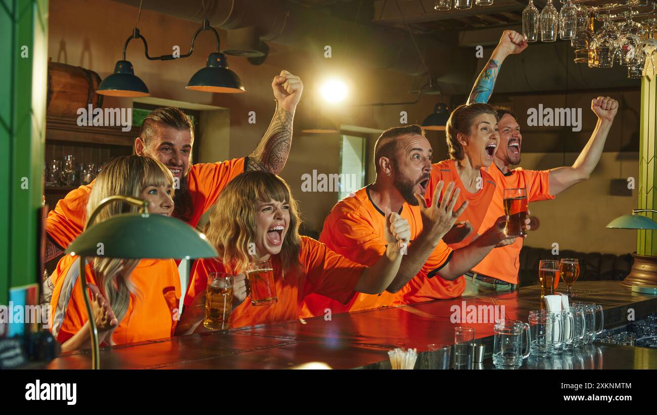 Group of football fans in orange shirts cheer loudly at bar raising ...
