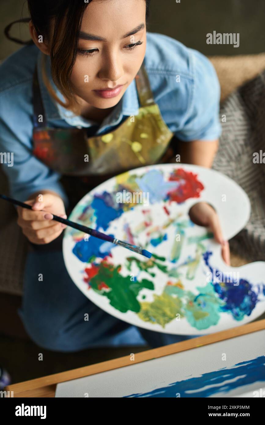 A young Asian woman in an apron holds a palette of paints ...