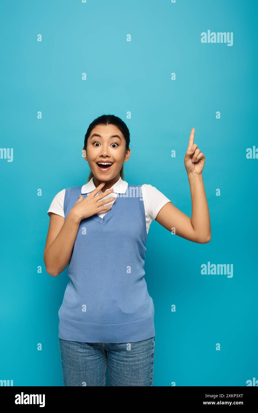 A young Asian woman in smart casual attire stands against a blue background, smiling and pointing upwards. Stock Photo