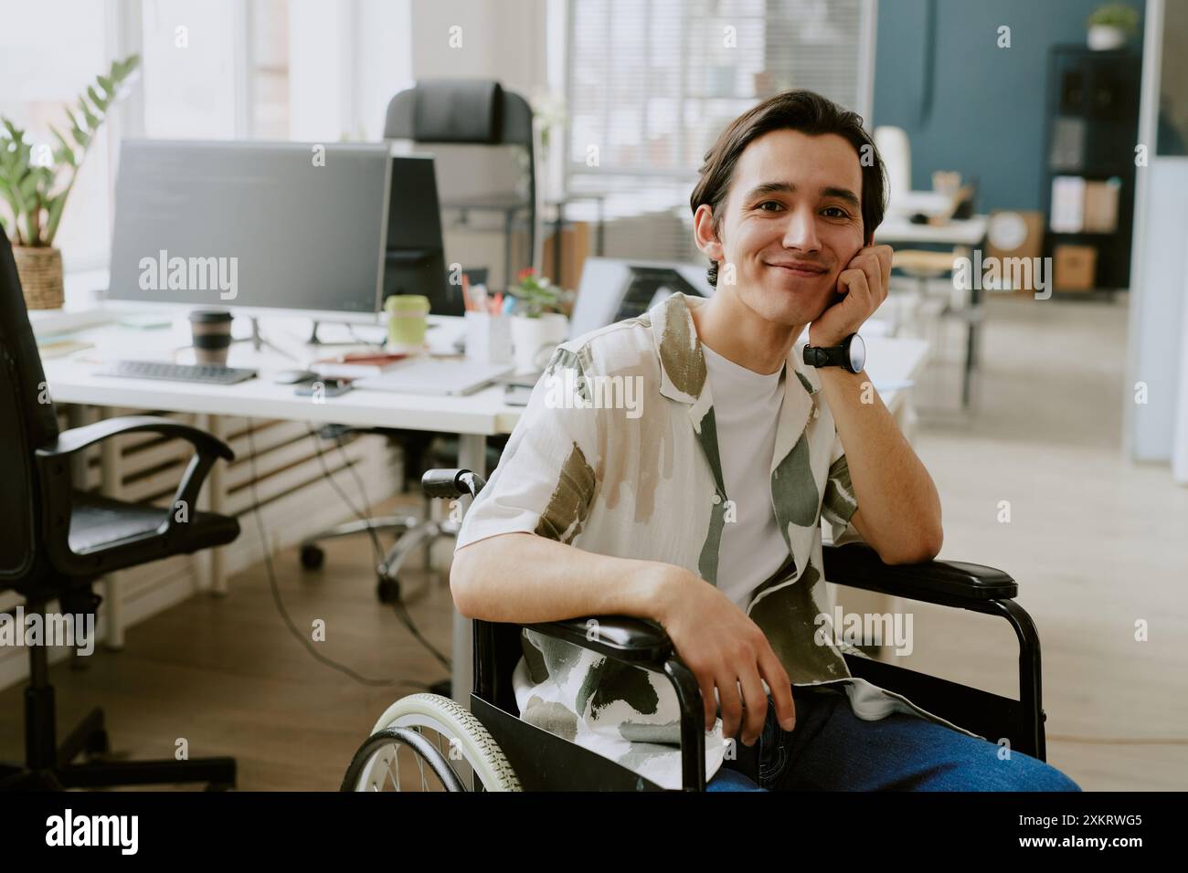 Portrait of smiling Caucasian IT specialist with disability looking at camera while his head based on hand, he in office of modern high-end IT company Stock Photo