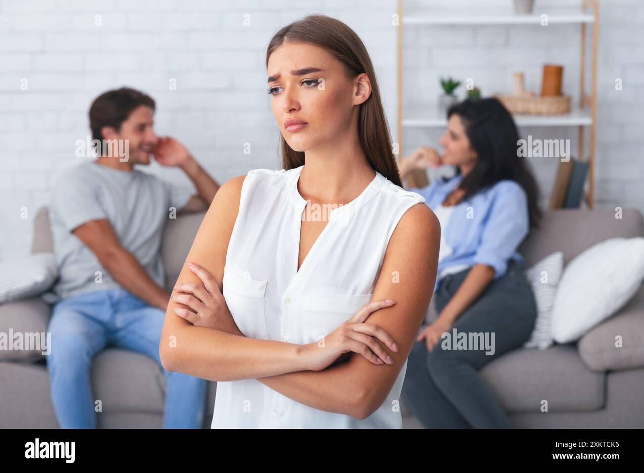 Jealousy Concept. Offended Girlfriend Standing Crossing Hands While Her Boyfriend Flirting With Girl Sitting On Sofa Indoor. Shallow Depth Stock Photo