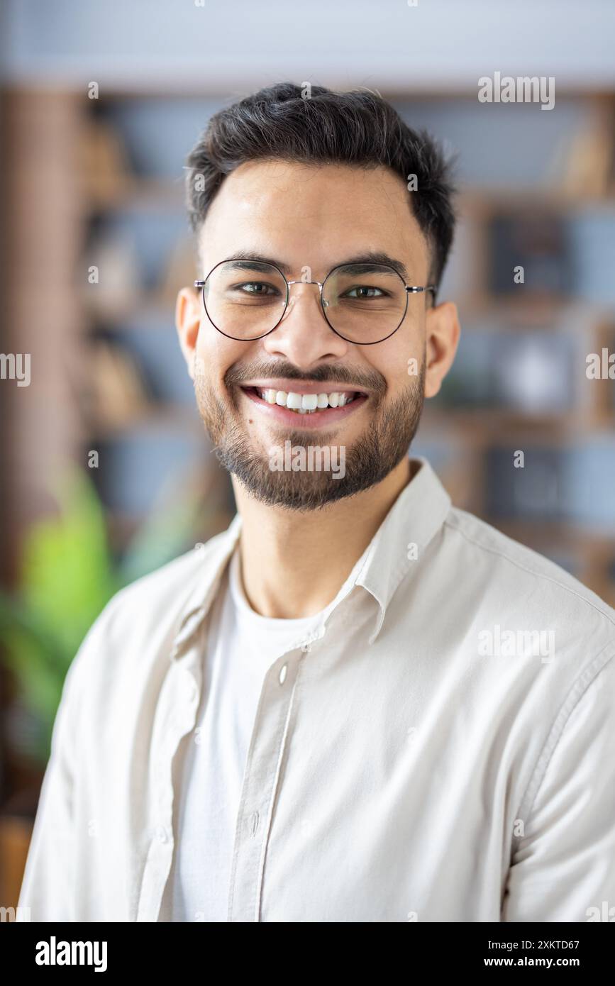 Smiling young man with glasses and beard wearing casual clothes indoors. Happy and confident expression, bright indoor setting, blurred background with bookshelves. Concept of positivity, youth, confidence, and casual style. Stock Photo