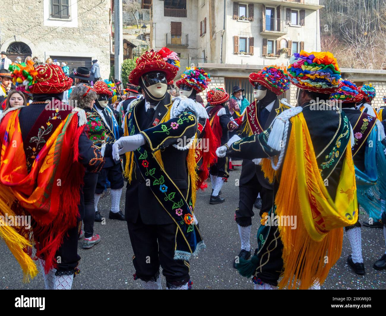 Balari dancing in Bagolino streets during Carnival, wearing the traditional costume with white knitted socks, black dress, colorful shawl over the back, face covered in a ivory and black mask, and head under a felt hat covered in red ribbon, with gold jewelry and multicolored ribbons forming a bow Stock Photo