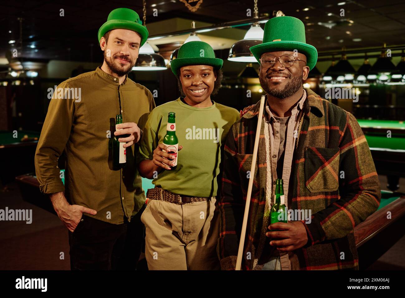 Three happy young friends in green hats and casual attire holding bottles of beer and looking at camera while standing in game room or pub Stock Photo