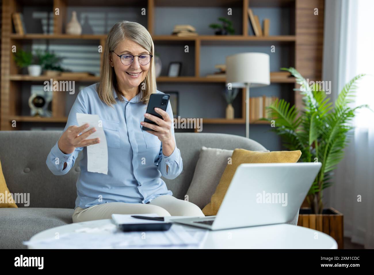 Older woman sitting on sofa using smartphone and holding receipt. Managing household budget, paying utilities and rent payments online. Happy and relaxed at home. Stock Photo