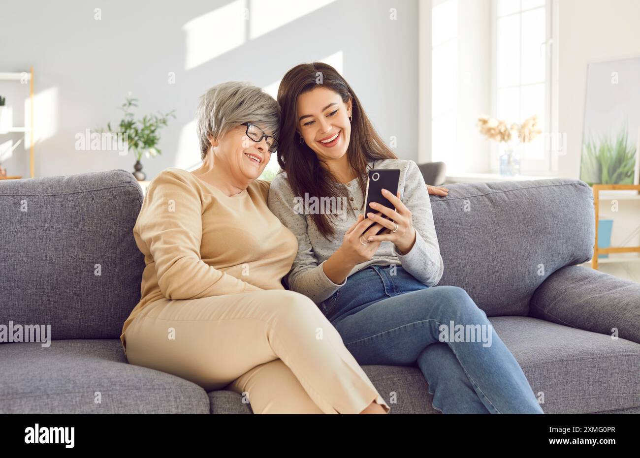 Adult woman using mobile phone with senior mother sitting at home and having online video call. Stock Photo