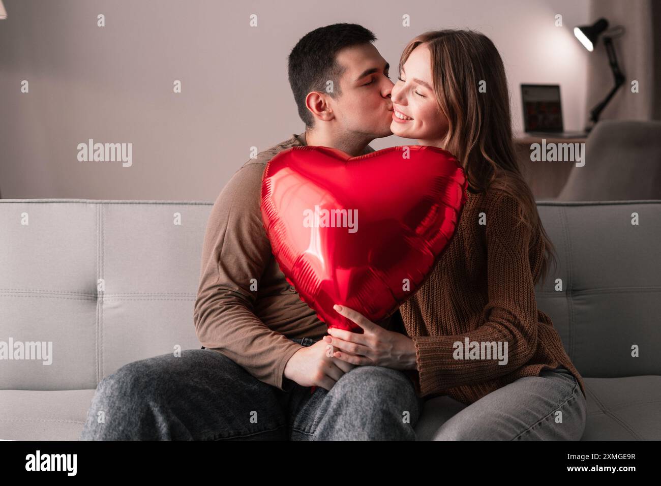 Romantic couple kissing on the sofa holding a red heart-shaped balloon ...