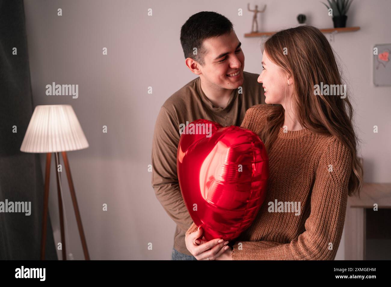Happy young couple in love with red heart-shaped balloon in living room ...