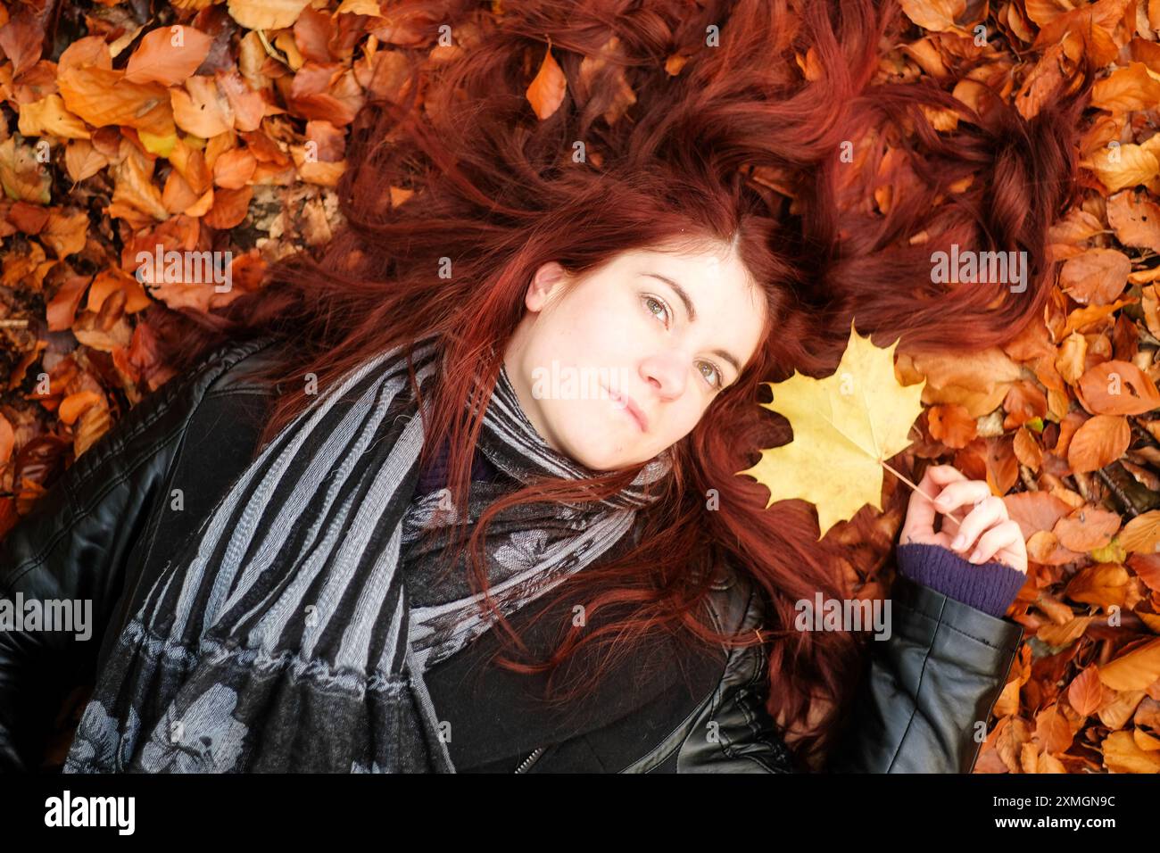 A woman with long red hair lies on a bed of autumn leaves, holding a ...
