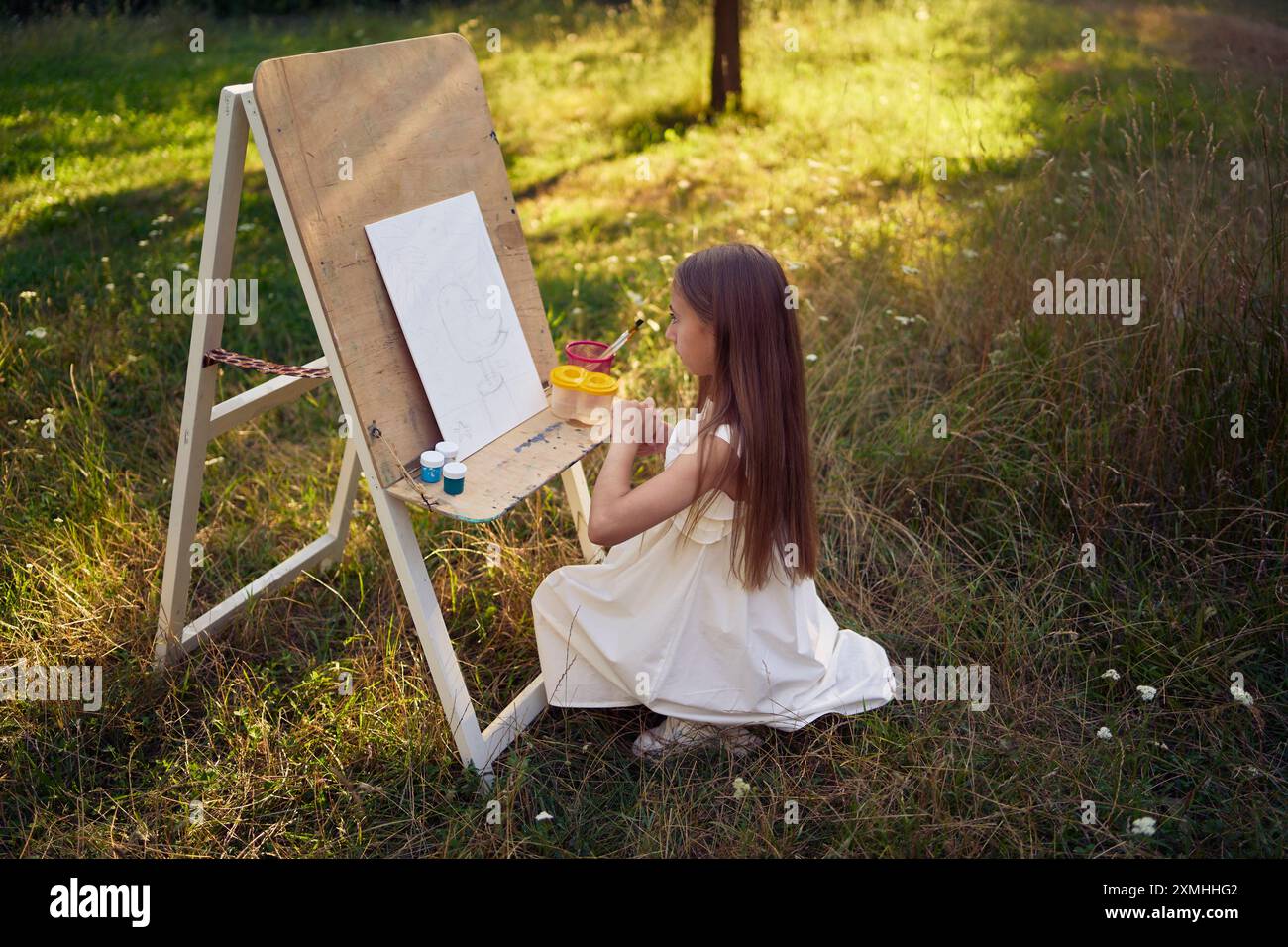 a pre-teen girl in a summer dress paints a picture in the garden on an ...