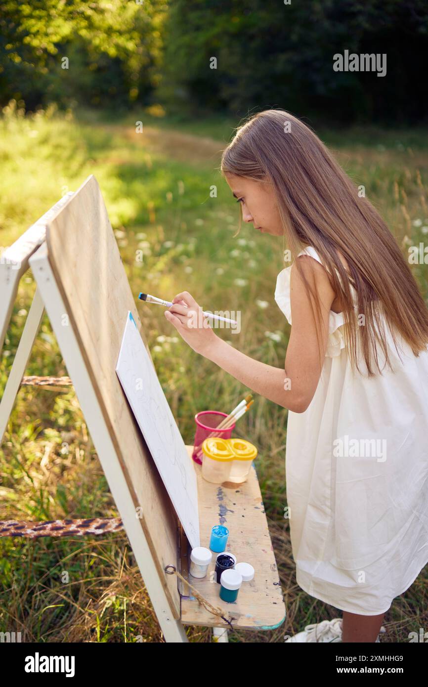 a pre-teen girl in a summer dress paints a picture in the garden on an ...