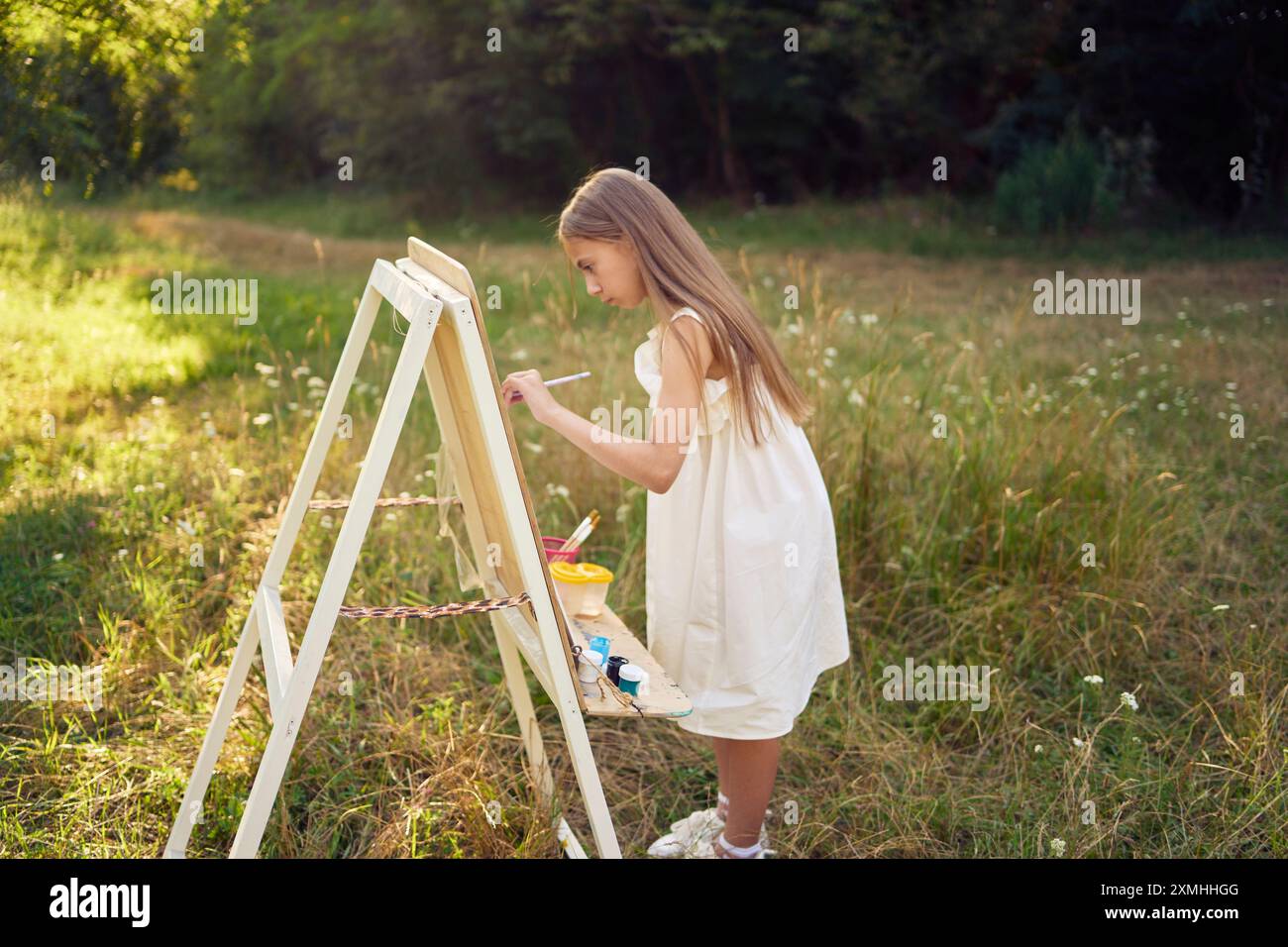 a pre-teen girl in a summer dress paints a picture in the garden on an ...