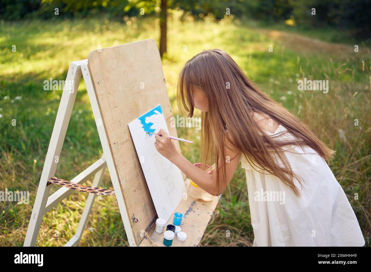 a pre-teen girl in a summer dress paints a picture in the garden on an ...