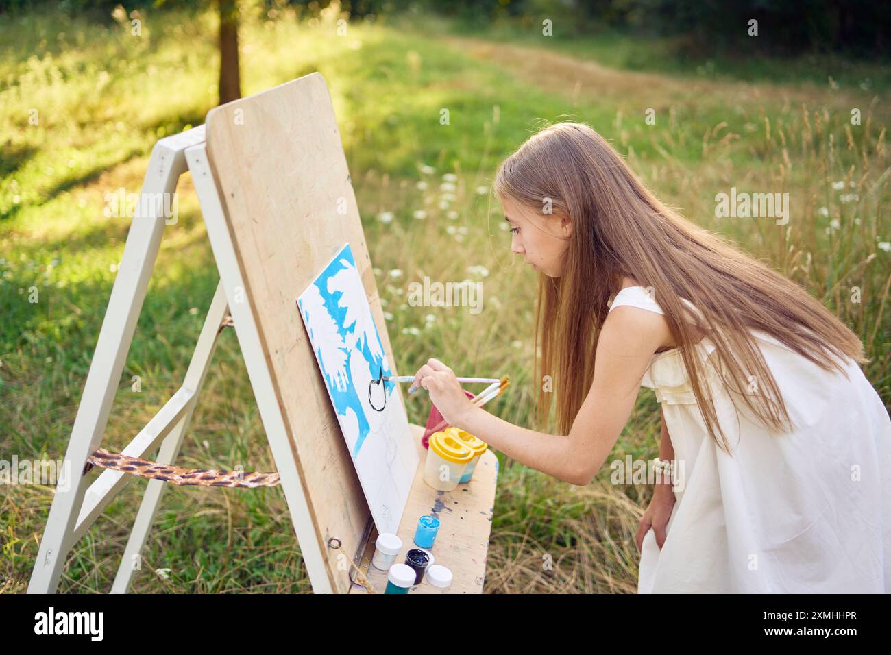 pre-teen girl in a summer dress paints a picture in the garden on an ...