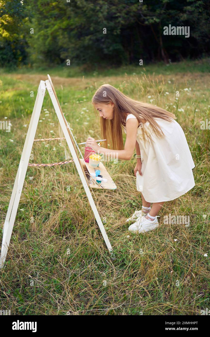 pre-teen girl in a summer dress paints a picture in the garden on an ...