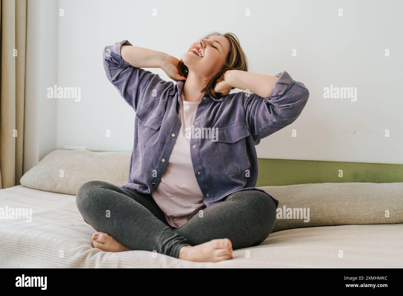 Happy plus-size woman sitting cross-legged on bed, stretching with a ...