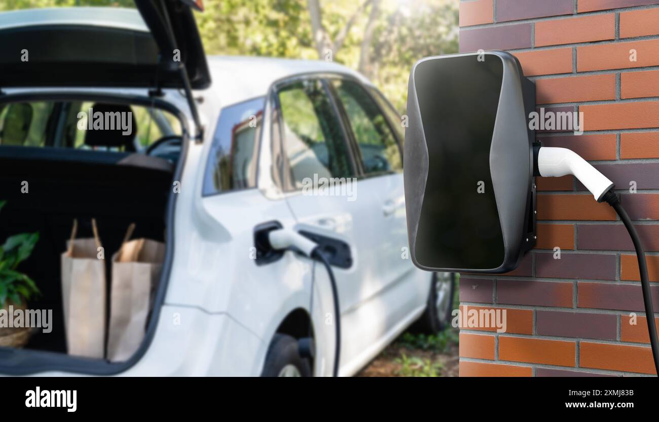 Charging electric car next to house with charging station on the wall. Stock Photo