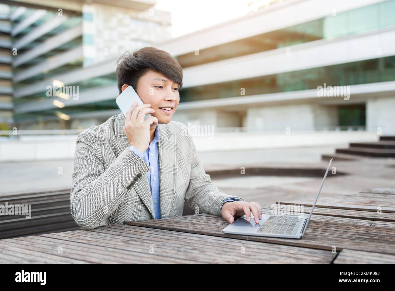 Young Man Talking on Phone, Using Laptop Outside Modern Building Stock Photo