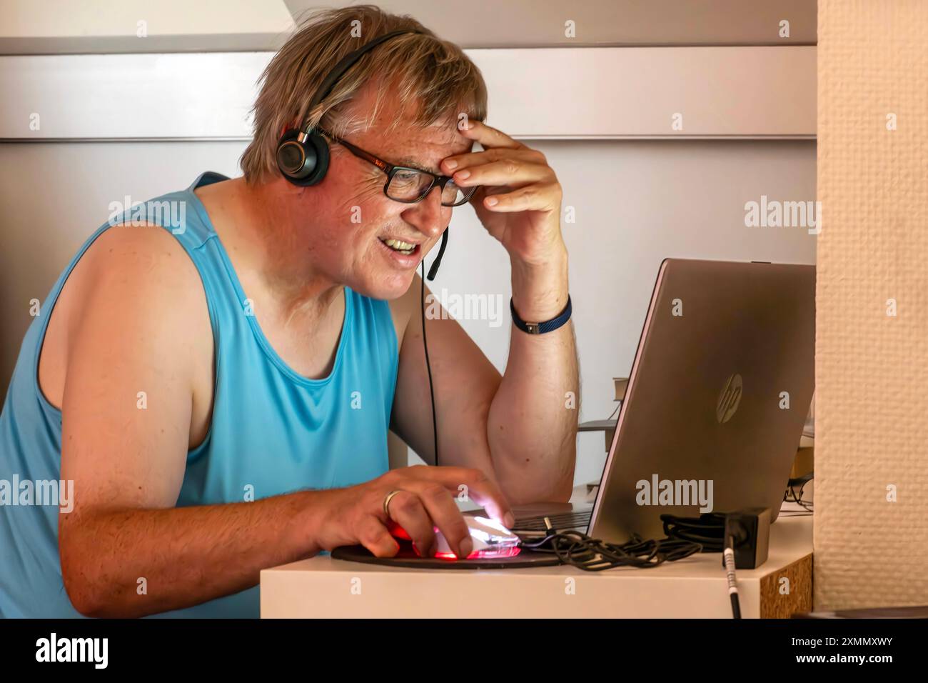 Mann schwitzt im Home Office, leidet unter Hitze in der Dachwohnung, München, Ende Juli 2024 Deutschland, München, Juli 2024, Mann schwitzt im Home Office, leidet unter Hitze in der Dachwohnung, sitzt unter der Dachschräge, sitzt im Unterhemd im Büro, er telefoniert und arbeitet am Computer, trägt ein Headset, das Notebook vor sich auf dem kleinen Schreibtisch, unpassende Kleidung für Videokonferenzen, privat, Arbeitszeit, Brille, Wohnung, Bayern, *** Man sweats in home office, suffers from heat in attic apartment, Munich, end of July 2024 Germany, Munich, July 2024, Man sweats in home office, Stock Photo