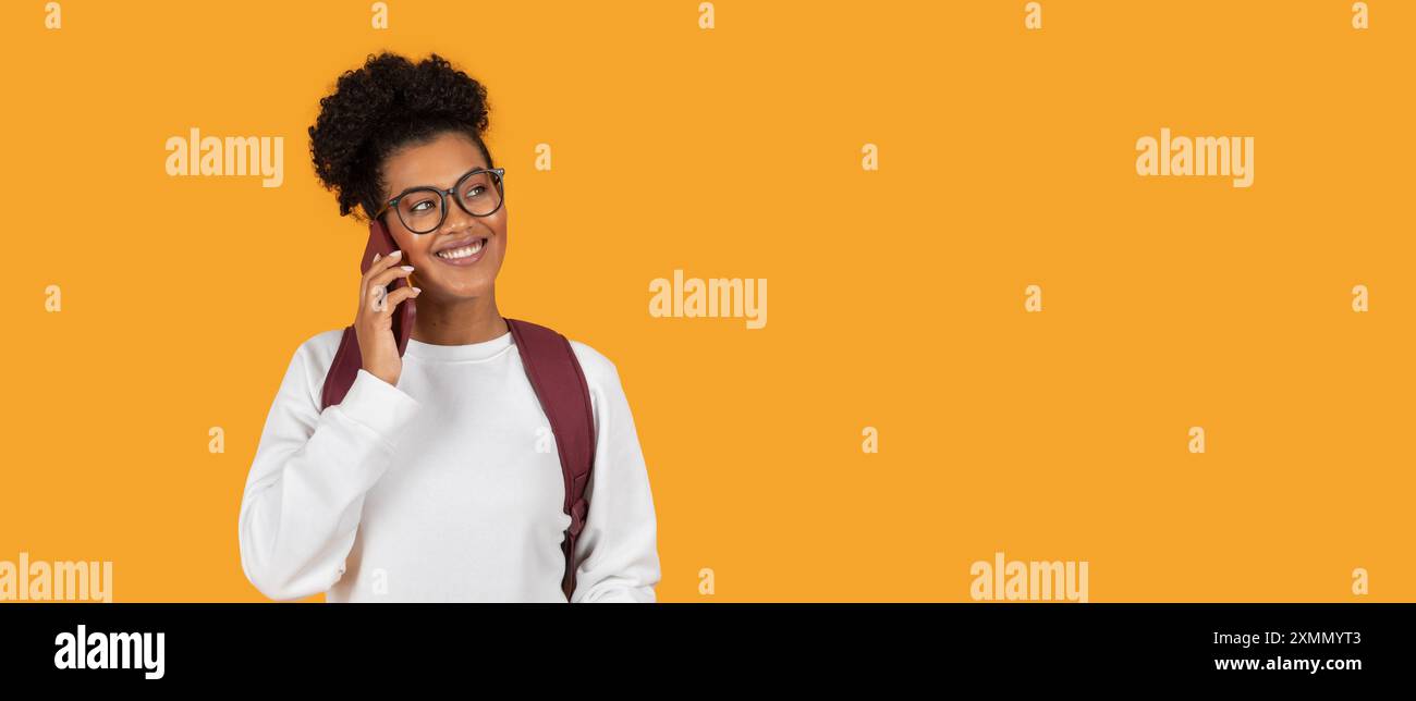 Smiling Young Woman Talking On Phone With Backpack Against Yellow Stock Photo