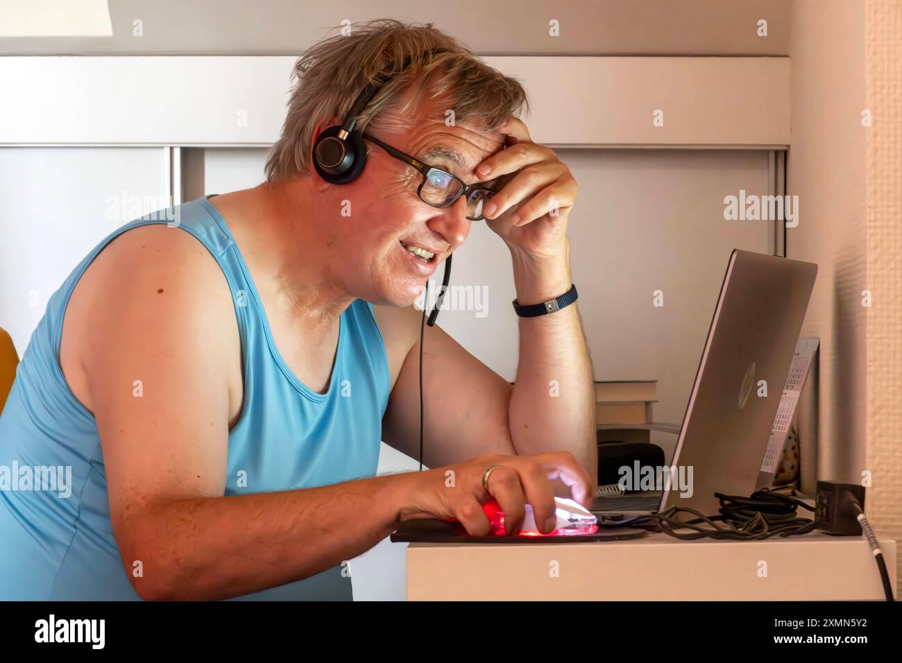 Älterer Mann schwitzt im Home Office, leidet unter Hitze in der Dachwohnung, München, Ende Juli 2024 Deutschland, München, Juli 2024, Mann schwitzt im Home Office, leidet unter Hitze in der Dachwohnung, sitzt unter der Dachschräge, sitzt im Unterhemd im Büro, er telefoniert und arbeitet am Computer, trägt ein Headset, das Notebook vor sich auf dem kleinen Schreibtisch, unpassende Kleidung für Videokonferenzen, privat, Arbeitszeit, Brille, Wohnung, Bayern, *** Elderly man sweats in home office, suffers from heat in attic apartment, Munich, end of July 2024 Germany, Munich, July 2024, man sweats Stock Photo