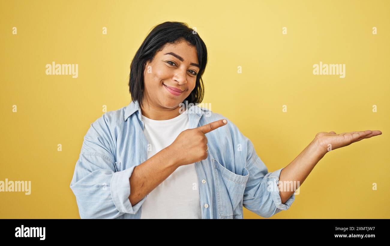A cheerful woman gestures pointing and presenting against a vibrant yellow background, embodying positivity and confidence. Stock Photo