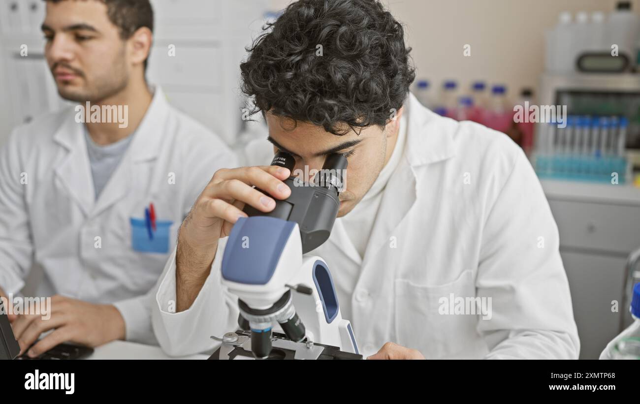 Two men in lab coats working in a laboratory, one using a microscope and the other taking notes. Stock Photo