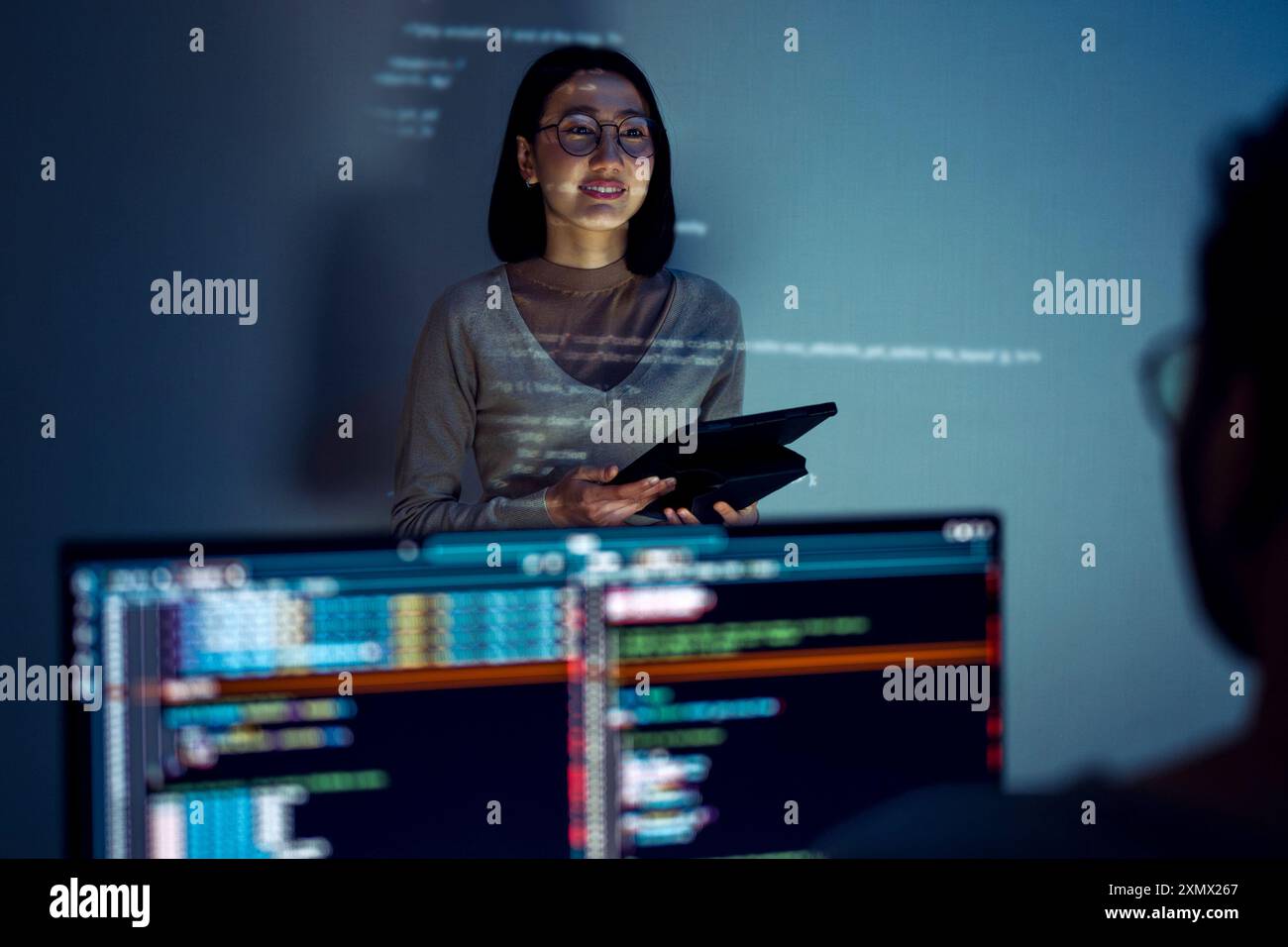 asian woman developers collaborating on a coding project, with one member presentation at code projected on the wall while the other works on multiple Stock Photo
