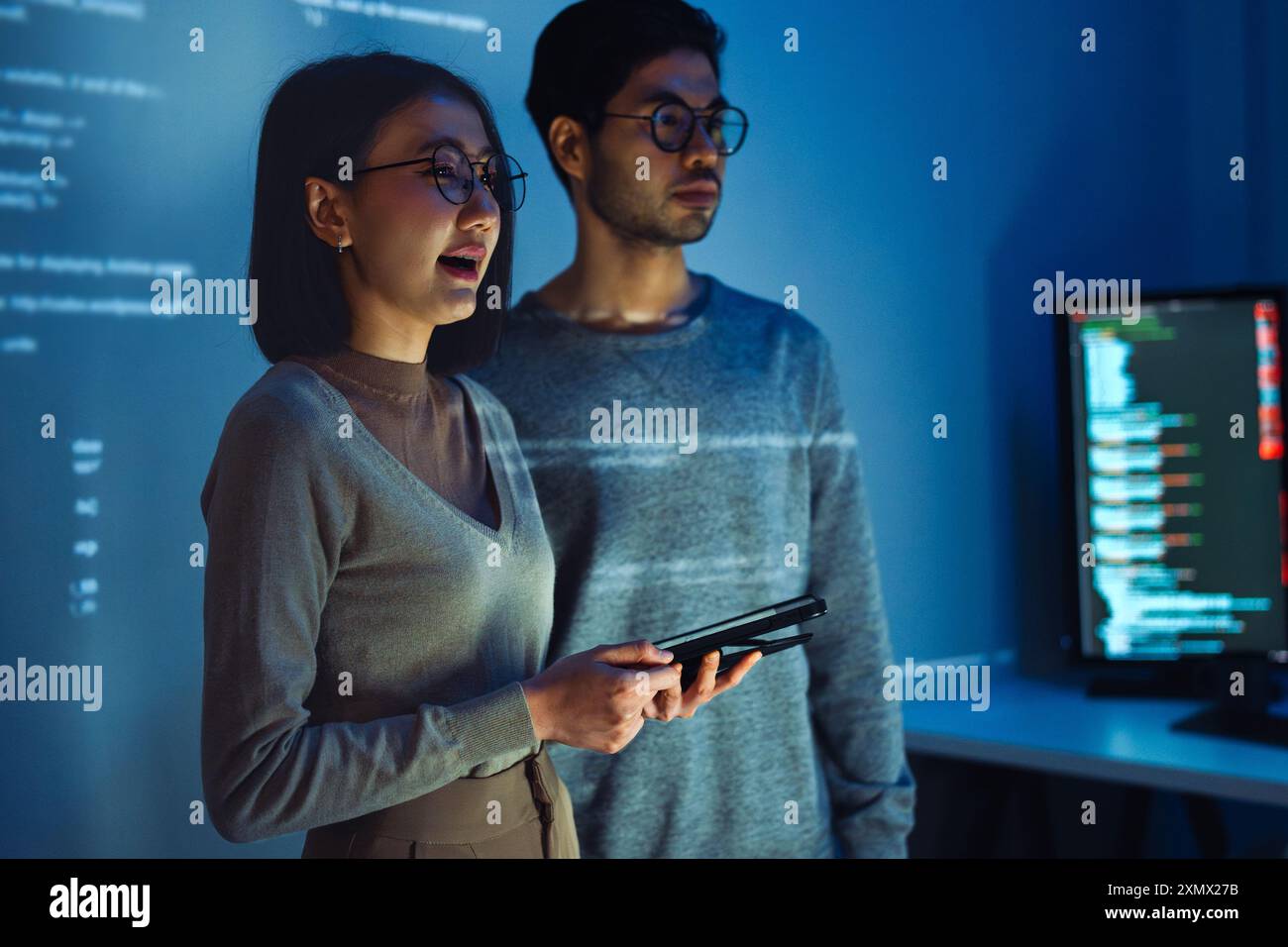 Asian Two software developers engaged in collaborative coding session, with code projected onto the wall and multiple computer screens displaying Stock Photo
