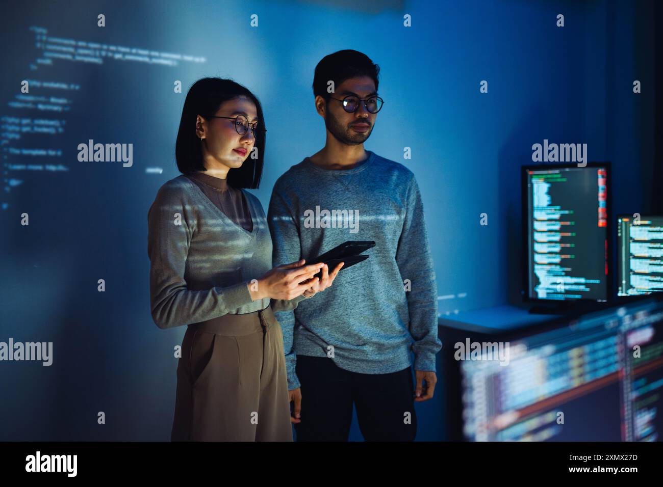 Asian Two software developers engaged in collaborative coding session, with code projected onto the wall and multiple computer screens displaying Stock Photo