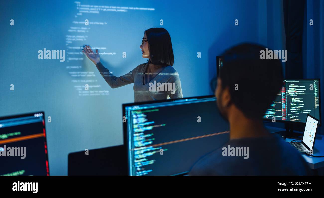 asian woman developers collaborating on a coding project, with one member pointing at code projected on the wall while the other works on multiple com Stock Photo