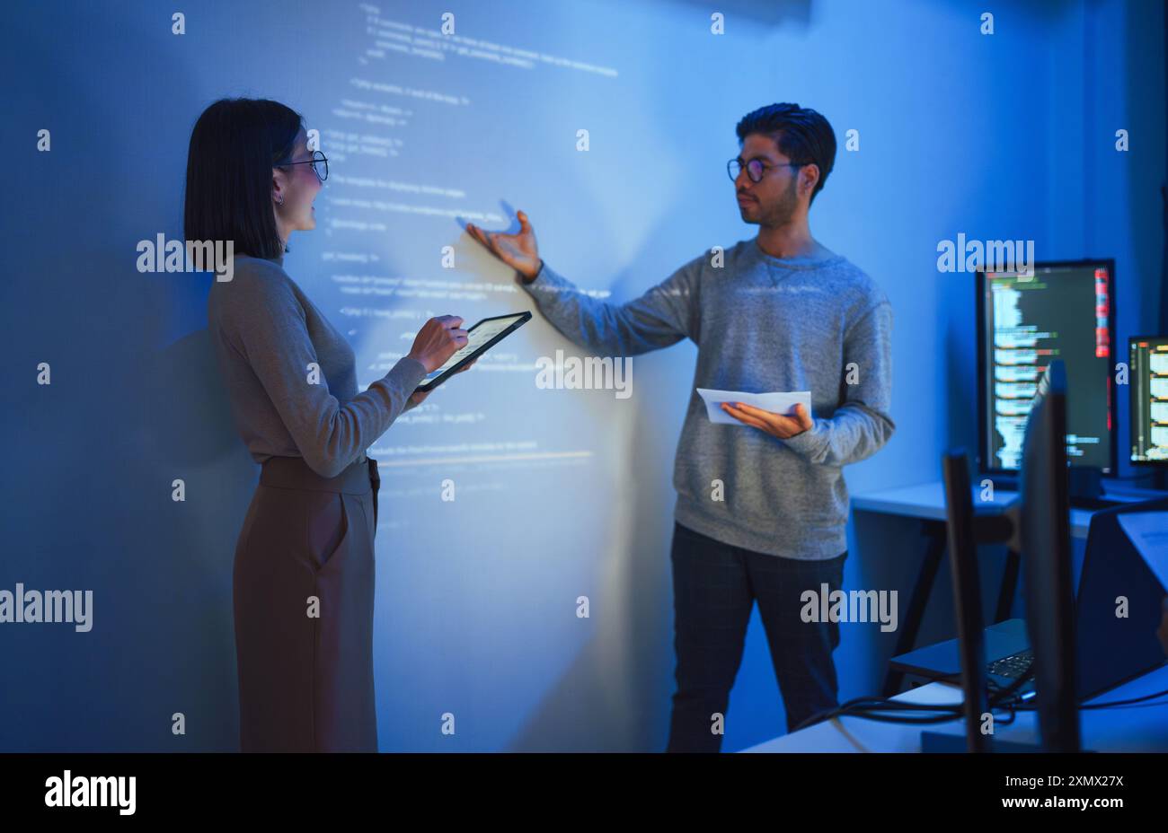 Asian Two software developers presentation in collaborative coding session, with code projected onto the wall and multiple computer screens displaying Stock Photo