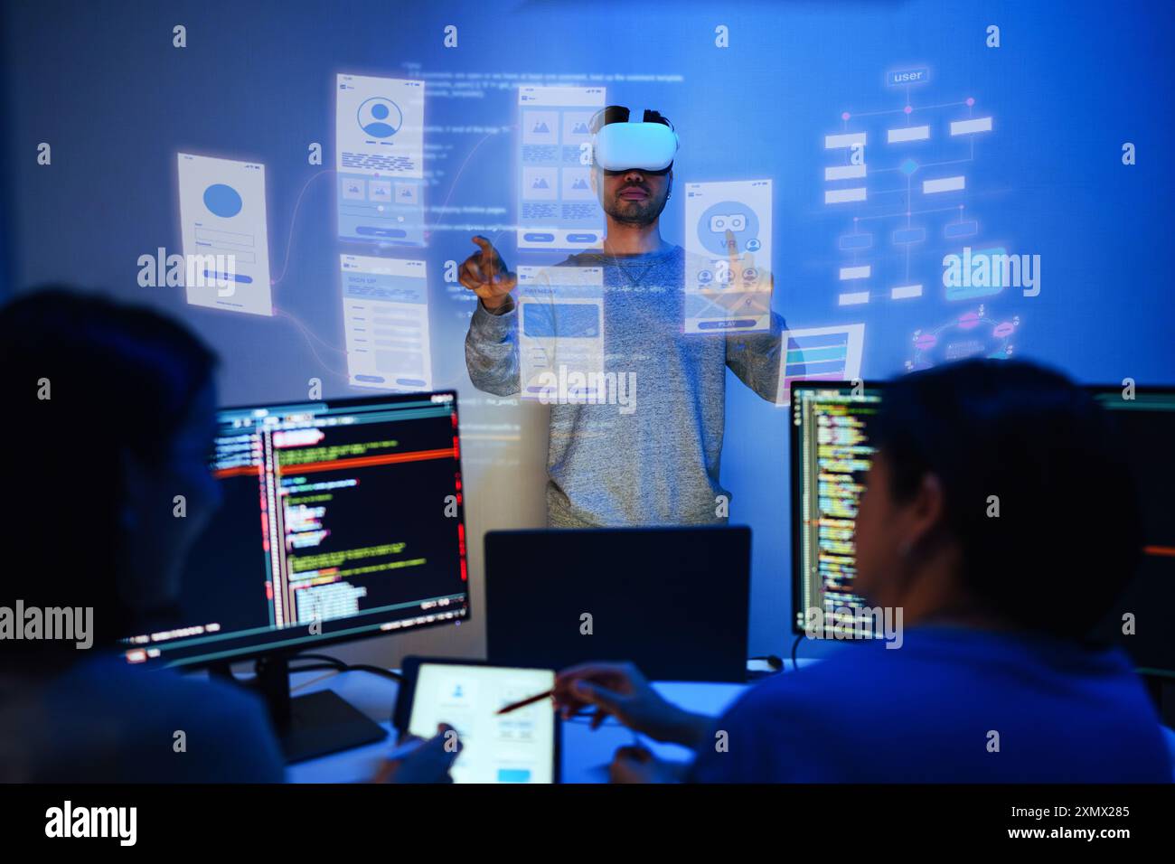 developer uses a VR headset to interact with projected interface designs, while colleagues discuss and code on screens at office Stock Photo