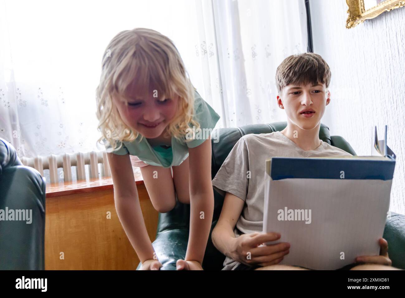 A young girl is peering over the back of a chair at a teenage boy reading a book. She is standing and bending over the back of the chair, while the bo Stock Photo