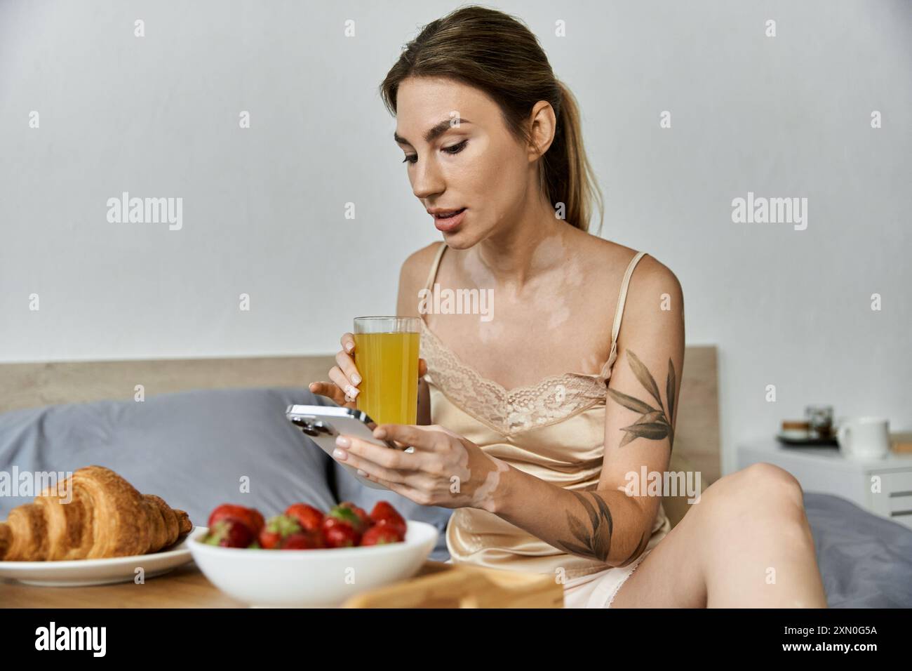 A young woman with vitiligo and a tattoo enjoys a leisurely morning in her apartment, sipping juice and scrolling through her phone. Stock Photo